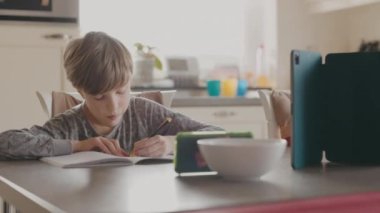 Mutfaktaki masada oturan çocuk telefona bakıyor ve not defterine yazıyor. Uzaklık Öğrenme.