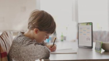 Mutfaktaki masada oturan çocuk telefona bakıyor ve not defterine yazıyor. Uzaklık Öğrenme.