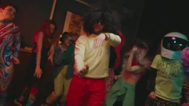 Gençler Partisi. Renkli elbiseli kızlar ve geyler zıplar ve dans eder. Uzun siyah saçlı ve ön planda başlıklı gay..