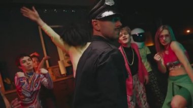 Gençler Partisi. Renkli elbiseli kızlar ve geyler. Polis üniformalı komik adam silahlı ve telsiz şakayla hareket ediyor..