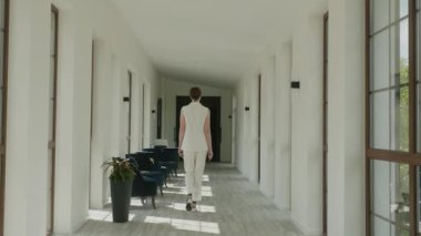 Beyaz Pantolon Giyen Model Otelin lobisinde yürüyor. Geniş pencereleri ve parlak sandalyeleri olan harika bir iç mekan. Uzun boylu esmer..