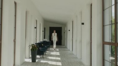 Beyaz Pantolon Giyen Model Otelin lobisinde yürüyor. Geniş pencereleri ve parlak sandalyeleri olan harika bir iç mekan. Uzun boylu esmer..