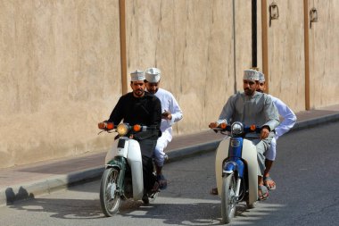 NIZWA, AL DAKHILIYAH, OMAN - 20 ARALIK 2024: Omani erkeklerinin geleneksel kıyafetleri ve motosikletleriyle Nizwa kalesinin önünde gezdikleri sokak sahnesi