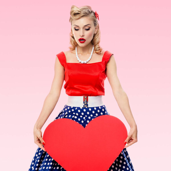 Woman holding heart symbol, dressed in pin-up style dress with polka dot, over pink background. Caucasian blond model posing in retro fashion and vintage concept studio shoot.