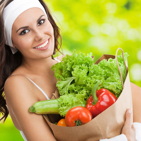 Portrait of happy smiling young beautiful woman in fitness wear holding grocery shopping bag with healthy vegetarian food, outdoors