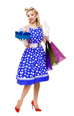 Puantiyeli mavi elbise giymiş, elinde hediye kutusu ve beyaz arka planda izole edilmiş alışveriş torbaları olan kadın portresi. Satış, tüketici, retro moda ve klasik konsept olarak poz veren beyaz sarışın bir model..
