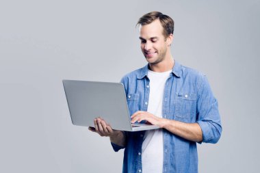 Mavi giysili mutlu gülümseyen genç adam portresi, gri arka planda dizüstü bilgisayarla çalışıyor.. 