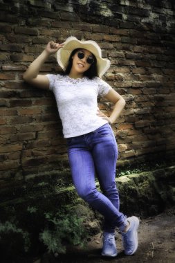 Beyaz dantelli bluzlu, kot pantolonlu ve hasır şapkalı genç bir kadın tuğla duvarın yanında poz veriyor. Rahat ve kendine güvenen bir yaz havası..