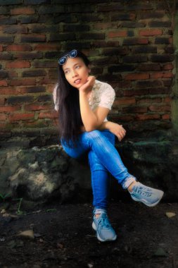 Beyaz dantelli bluz ve kot pantolon giymiş bir kadın eski bir duvara bağdaş kurmuş, çenesini eline dayayıp dinleniyor..