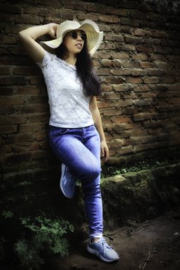Yazlık şapkalı, güneş gözlüklü ve kot pantolonlu bir kadın eskimiş bir duvara yaslanmış, refah hissi yayıyor..