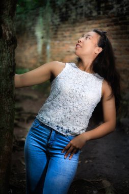 Açık renk bluzlu ve kot pantolonlu bir kadın ağacın yanında duruyor ve yukarı bakıyor, arka planda tuğla bir duvar var..