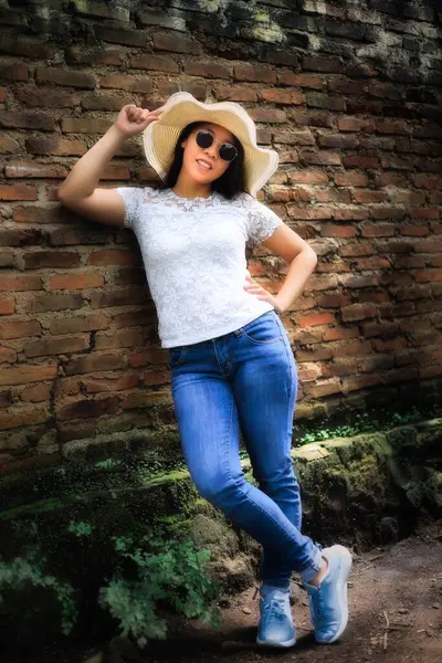 Beyaz dantelli bluzlu, kot pantolonlu ve hasır şapkalı genç bir kadın tuğla duvarın yanında poz veriyor. Rahat ve kendine güvenen bir yaz havası..