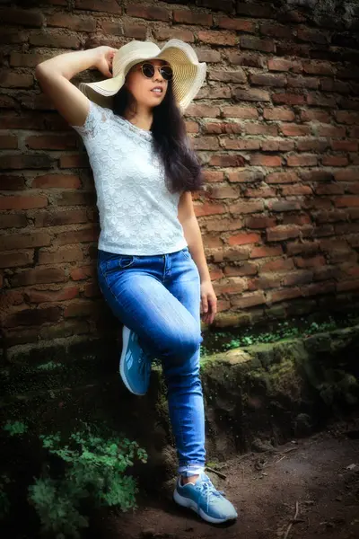 Yazlık şapkalı, güneş gözlüklü ve kot pantolonlu bir kadın eskimiş bir duvara yaslanmış, refah hissi yayıyor..