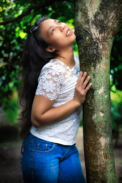 Beyaz dantelli bluz ve kotlu bir kadın ağaca sarılıyor ve hayal kurarak ormana bakıyor..