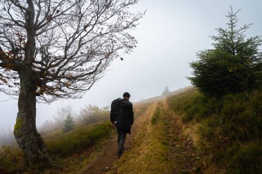 Sırt çantalı bir adam patika boyunca yürüyor, dağın tepesinde, sisli, bulutlu. Eğlence için. Seçici odak, doğa