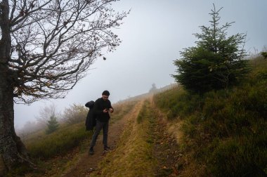 Sırt çantalı bir adam patika boyunca yürüyor, dağın tepesinde, sisli, bulutlu. Eğlence için. Seçici odak, doğa