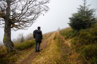 Sırt çantalı bir adam patika boyunca yürüyor, dağın tepesinde, sisli, bulutlu. Eğlence için. Seçici odak, doğa