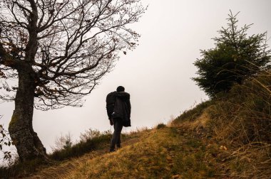 Sırt çantalı bir adam patika boyunca yürüyor, dağın tepesinde, sisli, bulutlu. Eğlence için. Seçici odak, doğa
