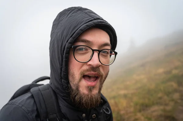 Bearded man in the mountains. A traveler with a backpack and glasses. Glasses and a beard are on his face. Age 32-35 years, fog. a trip, a trip, a break from the city and depression. Up close