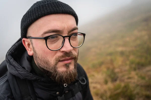 Bearded man in the mountains. A traveler with a backpack and glasses. Glasses and a beard are on his face. Age 32-35 years, fog. a trip, a trip, a break from the city and depression. Up close