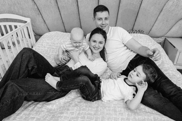 Young, emotional, cheerful, big family, mom, dad and sons lie on a big bed and look at the camera.