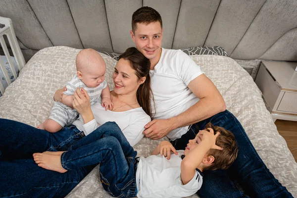 Young, emotional, cheerful, big family, mom, dad and sons lie on a big bed and look at the camera.