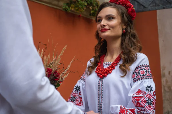 A happy young couple in love, a family walking through the old city of Lviv in Ukrainian embroidered shirts, holding hands on a red-orange background. Young people hug in the old town of Lviv