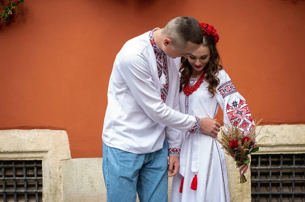 A happy young couple in love, a family walking through the old city of Lviv in Ukrainian embroidered shirts, holding hands on a red-orange background. Young people hug in the old town of Lviv