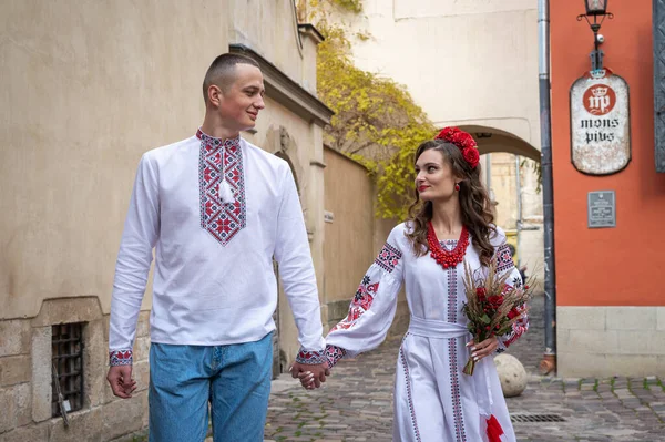 Happy young couple in love, family walking in the old city of Lviv in traditional Ukrainian shirts, holding hands on a red-orange background. Young people hug in the old town of Lviv