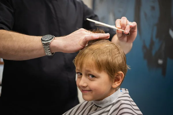 Happy cute fair-haired preschool boy getting a haircut. Children\'s hairdresser with scissors and comb cuts a little boy\'s hair in a room with a loft interior