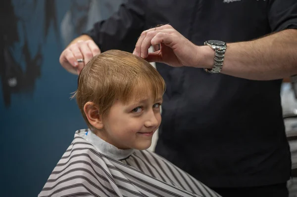 Happy cute fair-haired preschool boy getting a haircut. Children\'s hairdresser with scissors and comb cuts a little boy\'s hair in a room with a loft interior