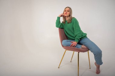 Young, cute, emotional, chic girl sitting on a chair, flirtatious mood, empty space, wearing jeans and green sweater on isolated white background, place for text. Full length photo