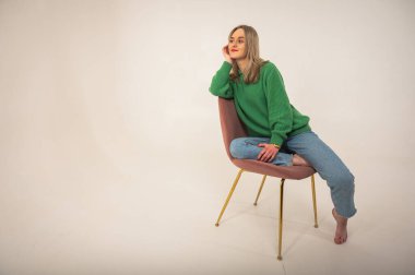 Young, cute, emotional, chic girl sitting on a chair, flirtatious mood, empty space, wearing jeans and green sweater on isolated white background, place for text. Full length photo