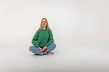 Young, cute, emotional, chic girl sitting on the floor in a flirtatious mood, hand gestures, empty space, wearing jeans and a green sweater on an isolated white background, place for text