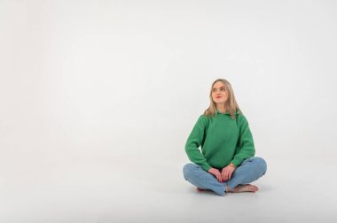 Young, cute, emotional, chic girl sitting on the floor in a flirtatious mood, hand gestures, empty space, wearing jeans and a green sweater on an isolated white background, place for text