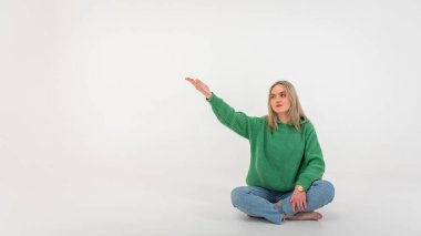 portrait of a young emotional girl flirtatiously stretching her arms to the side, gesturing with her hands, empty space, wearing jeans and a green sweater on an isolated white background