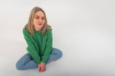 Young, cute, emotional, chic girl sitting on the floor in a flirtatious mood, hand gestures, empty space, wearing jeans and a green sweater on an isolated white background, place for text
