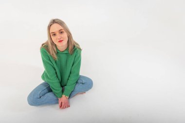 Young, cute, emotional, chic girl sitting on the floor in a flirtatious mood, hand gestures, empty space, wearing jeans and a green sweater on an isolated white background, place for text