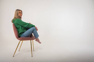Young, cute, emotional, chic girl sitting on a chair, flirtatious mood, empty space, wearing jeans and green sweater on isolated white background, place for text. Full length photo