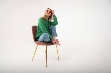 Young, cute, emotional, chic girl sitting on a chair, flirtatious mood, empty space, wearing jeans and green sweater on isolated white background, place for text. Full length photo