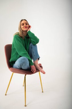 portrait of young, emotional girl sitting on floor in flirty mood, gesturing with hands, empty space, wearing jeans and green sweater on isolated white background, place for text 