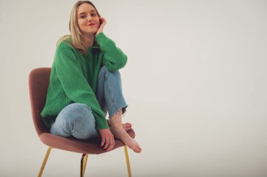 Young, cute, emotional, chic girl sitting on a chair, flirtatious mood, empty space, wearing jeans and green sweater on isolated white background, place for text. Full length photo
