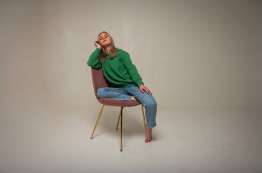 Young, cute, emotional, chic girl sitting on a chair, flirtatious mood, empty space, wearing jeans and green sweater on isolated white background, place for text. Full length photo