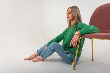 portrait of a young emotional girl sitting on the floor near a chair in a flirtatious mood, gesturing with her hands, empty space, wearing jeans and a green sweater n, space for text