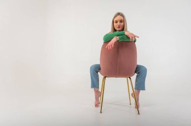 portrait of a young, emotional girl sitting on a chair in a flirtatious mood, gesturing with her hands, empty space, wearing jeans and a green sweater on an isolated white background, space for text