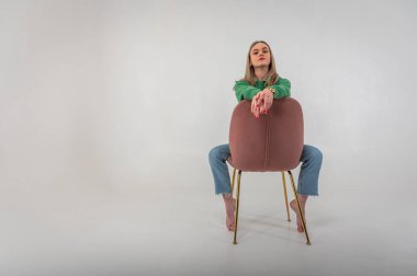 portrait of a young, emotional girl sitting on a chair in a flirtatious mood, gesturing with her hands, empty space, wearing jeans and a green sweater on an isolated white background, space for text