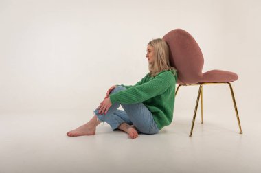 portrait of a young emotional girl sitting on the floor near a chair in a flirtatious mood, gesturing with her hands, empty space, wearing jeans and a green sweater n, space for text