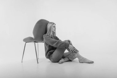 portrait of a young emotional girl sitting on the floor near a chair in a flirtatious mood, gesturing with her hands, empty space, wearing jeans and a green sweater n, space for text