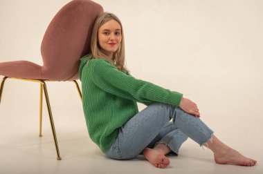 portrait of a young emotional girl sitting on the floor near a chair in a flirtatious mood, gesturing with her hands, empty space, wearing jeans and a green sweater n, space for text