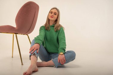portrait of a young emotional girl sitting on the floor near a chair in a flirtatious mood, gesturing with her hands, empty space, wearing jeans and a green sweater n, space for text